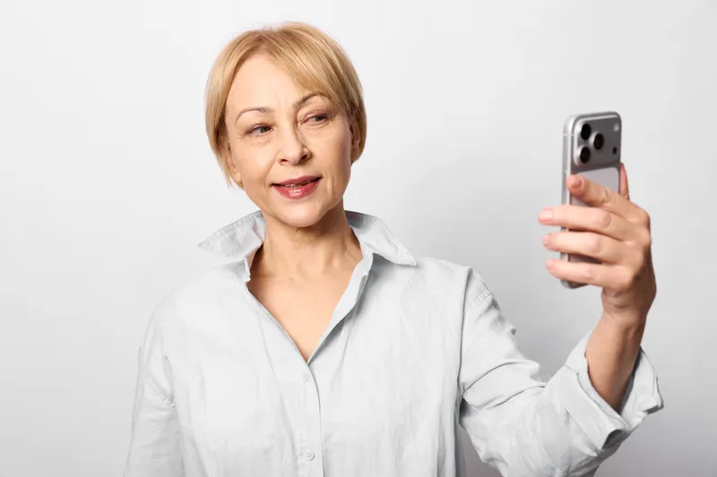 Woman selfie with smartphone, smiling portrait, casual shirt, white studio background, relaxed pose and soft light, modern communication and everyday self confidence