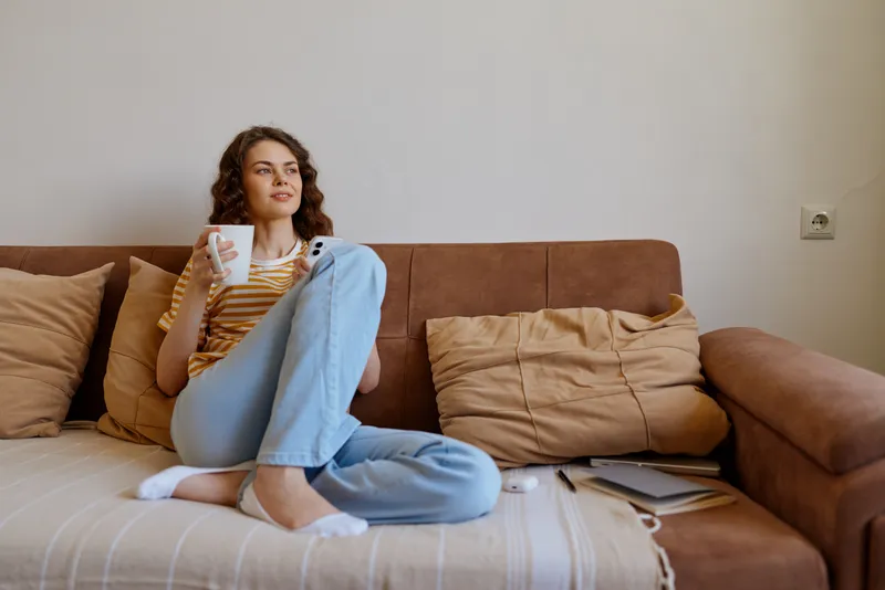 A woman sits on a sofa at home, wearing a striped top and blue jeans, barefoot, holding a mug and looking away. A cozy, casual moment featuring relaxed mornings and comfortable domestic mood.