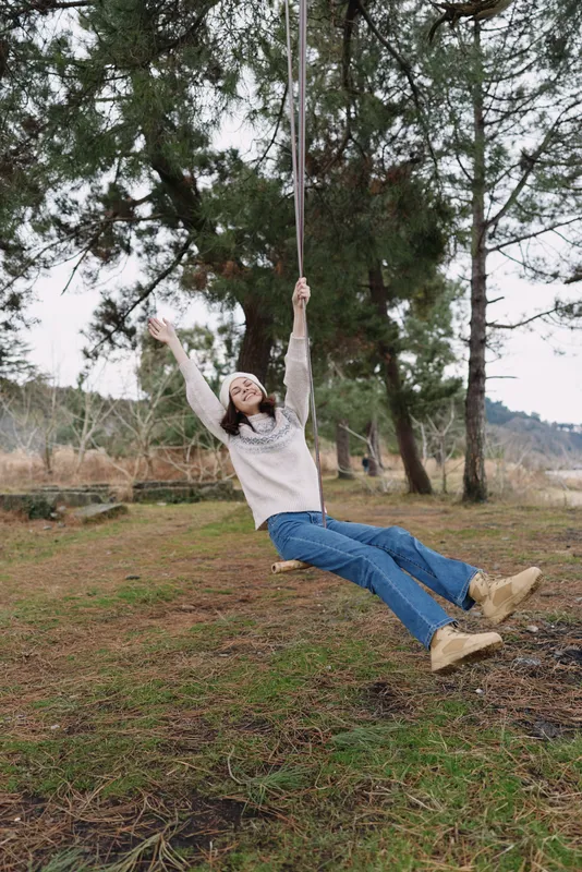 Woman on a swing outdoors in a park, arms raised, joyful moment among trees, casual attire, and open space, capturing carefree energy and fresh air during daytime.