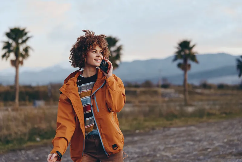 Woman lifestyle outdoors, curly hair, orange jacket, smiling, holding phone at ear, rural landscape with palm trees and distant mountains, warm sunlight