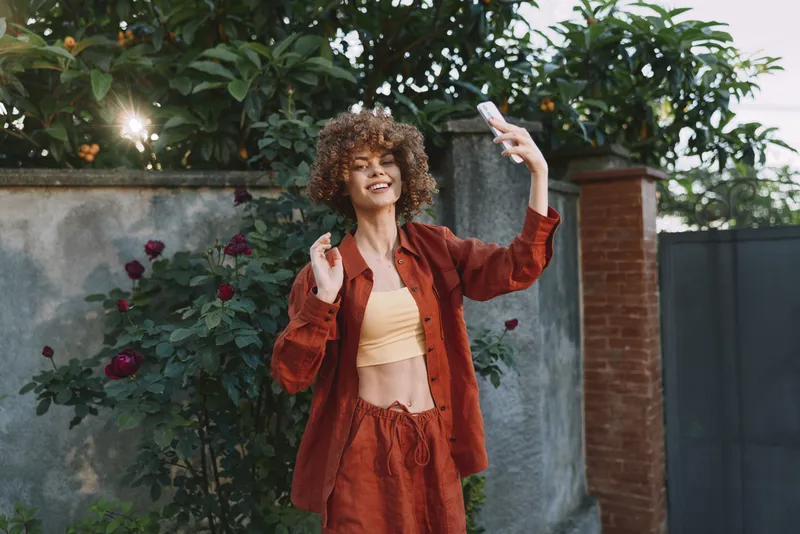 Cheerful woman outdoors, taking a selfie with a smartphone, sunlit moment, relaxed posture, warm rust tones, casual outfit, curly hair, lush greenery backdrop garden wall, soft breeze, natural colors