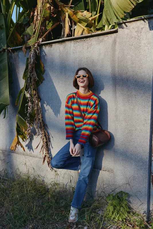 Young woman in a colorful striped sweater and sunglasses leaning wall by tropical leaves, smiling and laughing in casual outfit and jeans for travel lifestyle and street fashion.