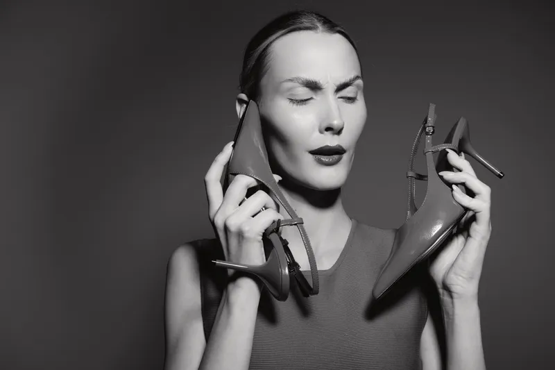 Caucasian woman with light skin holds two high heel shoes near face with closed eyes. Concept of fashion, style, and shoe choice in black and white photo.