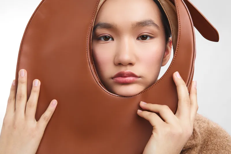 asian woman wears a large leather bag around her face, creating a surreal studio portrait with a soft, neutral backdrop and a calm, contemplative mood.