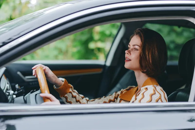 A woman sits behind the steering wheel of a car, focused on the road with a calm expression, casual sweater and natural light highlighting the interior.