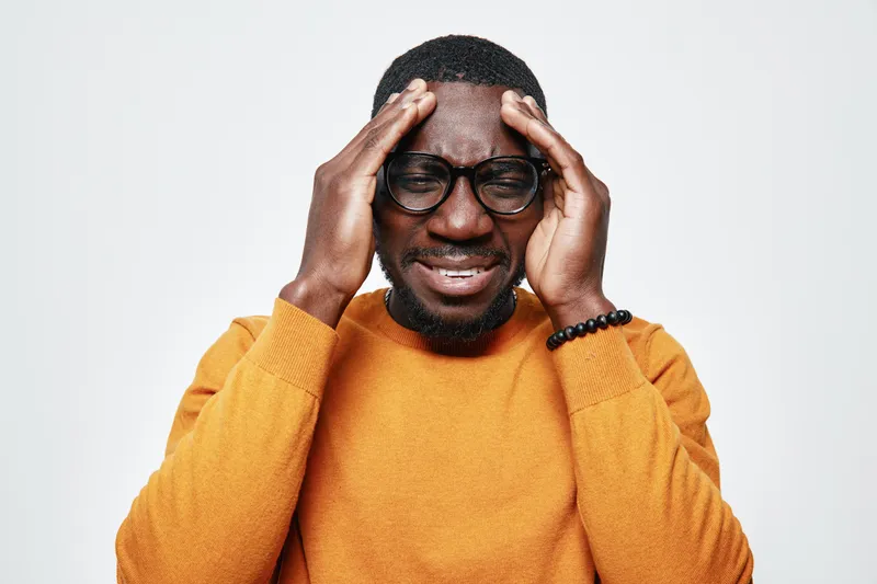 African man with dark skin and glasses wearing orange sweater holding head with pain or stress on white background. Male, portrait and discomfort with closed eyes and beard.