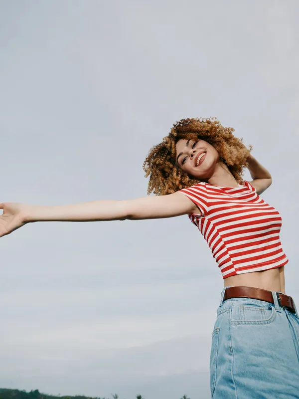 Joyful woman outdoors stretches arms wide in a red and white striped top, denim jeans, and brown belt, smiling under a clear blue sky.