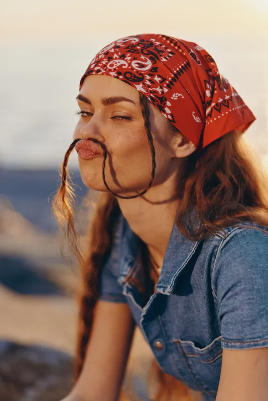Lifestyle woman in modern boho-western denim street style with playful expression and film color tones, wearing red bandana and braided hair outdoors.