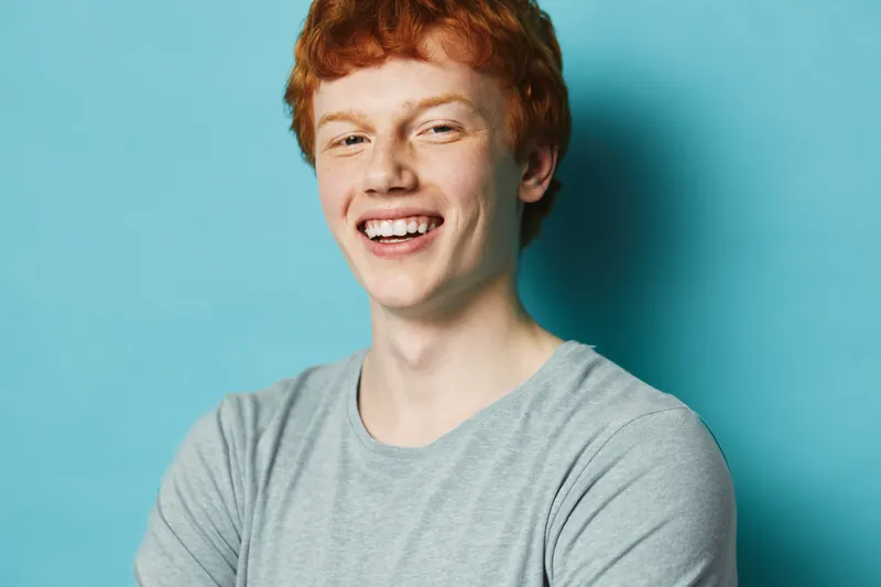 Young man with red hair and freckles smiling happily wearing casual grey t shirt standing against blue background in studio portrait. Positive emotion and confidence.