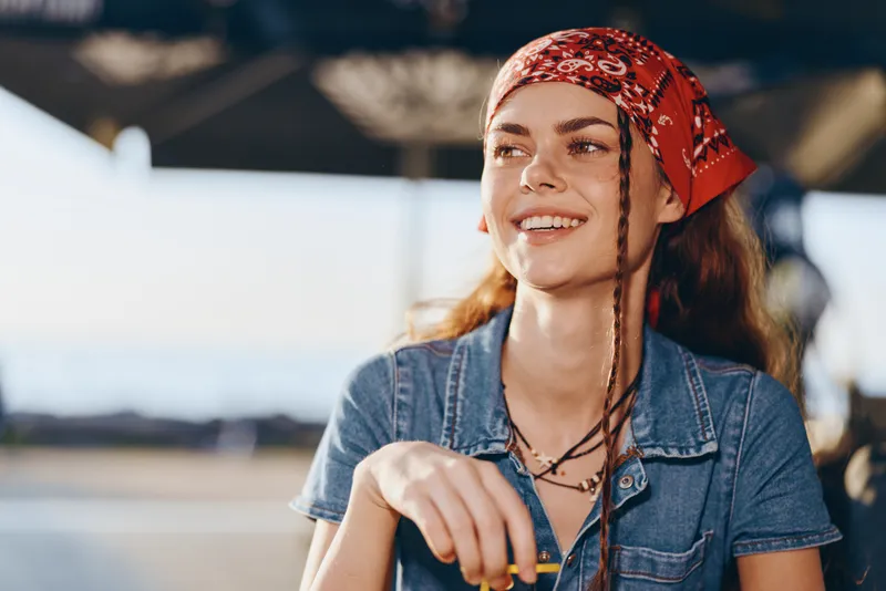 smiling woman with braided hair wearing red bandana and denim shirt outdoors in bright daylight with blurred background