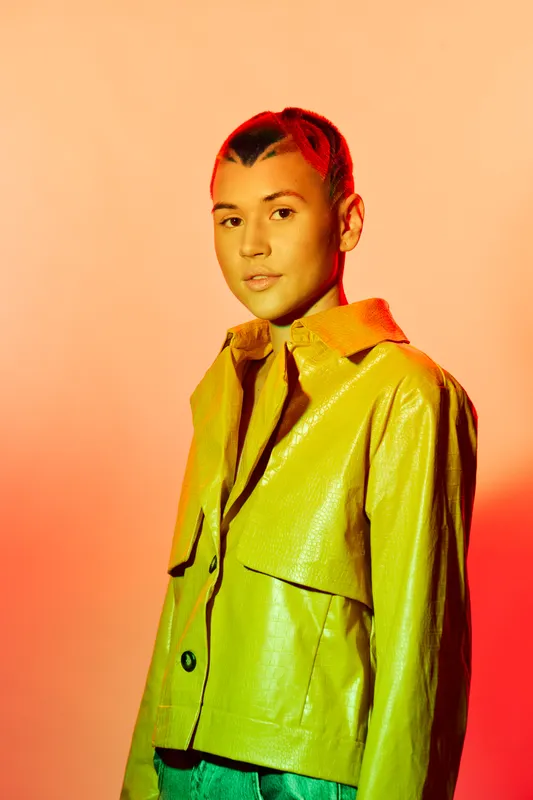 Young woman in a green leather jacket posing confidently against a vibrant orange gradient background, showcasing modern fashion and bright studio lighting effects.
