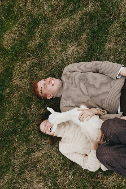 Young couple lies on grass smiling joyfully while holding a white baby goat in a cozy lifestyle outdoor setting during autumn season.