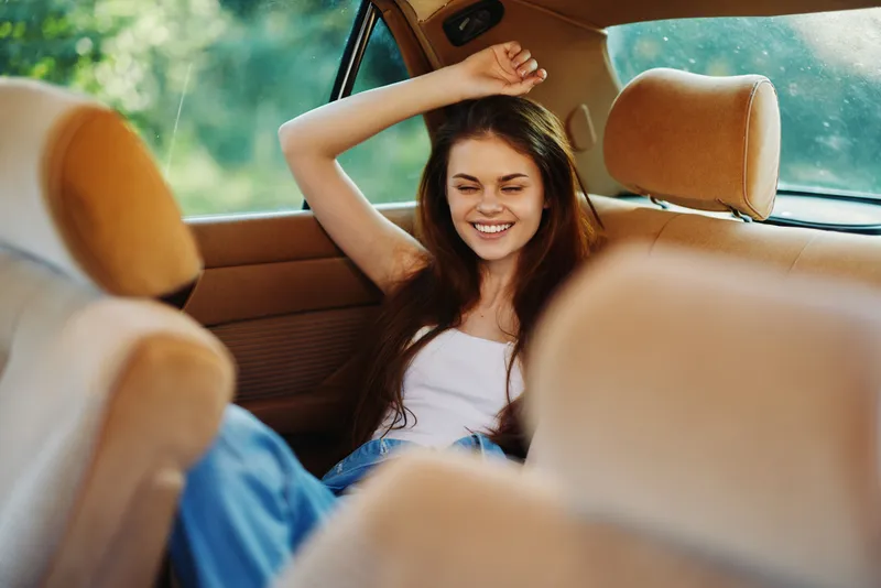 Happy young woman with long hair enjoying a carefree moment in the back seat of a car surrounded by nature, dressed casually in a tank top and jeans.