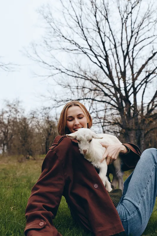 Woman outdoors in a casual jacket sits on the grass, cuddling a playful goat kid, a sunlit lifestyle moment highlighting friendship, calm nature, relaxed joy, and gentle affection