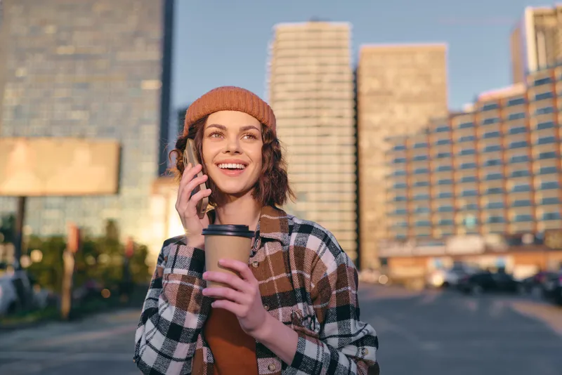 woman coffee phone smile urban beanie candid lifestyle portrait in golden hour glow, authenticity and mindful living captured with emotional storytelling in contemporary city street.
