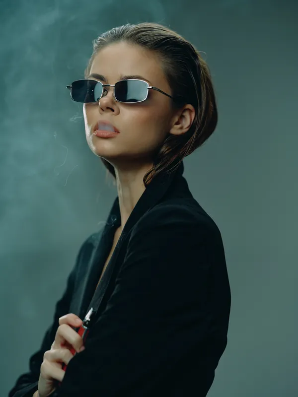 Stylish woman wearing sunglasses and black jacket exhaling smoke in moody indoor setting