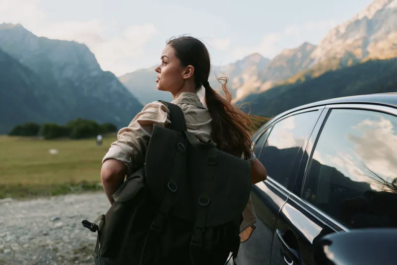 Young woman with backpack standing next to car in mountain landscape. Outdoor lifestyle, travel and adventure concept with natural light and scenic background.
