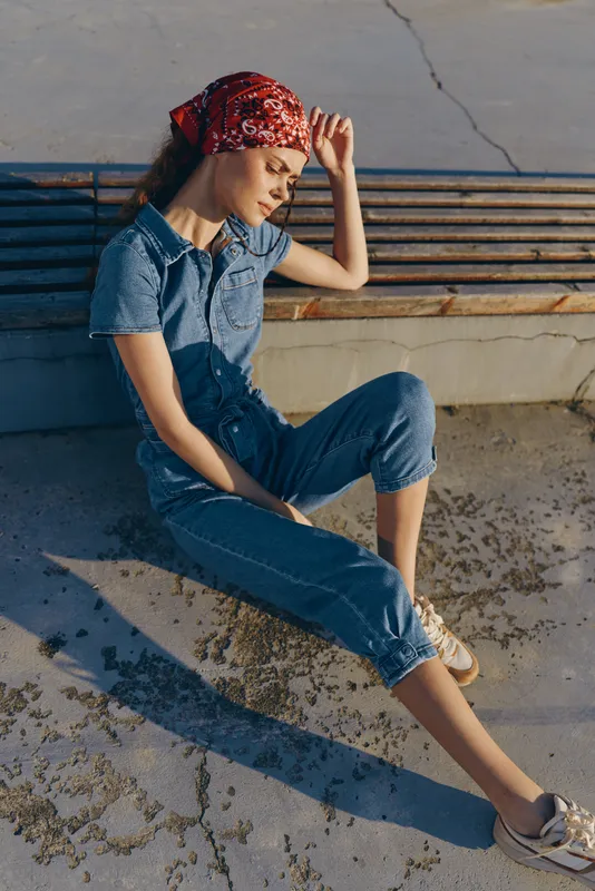 young woman sitting outdoors in casual denim jumpsuit and red bandana, relaxed mood, sunny day, urban setting, natural light, stylish outfit