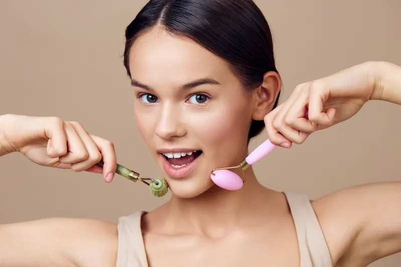 Young woman enjoying facial massage with green and pink rollers, showcasing a joyful expression against a neutral background, promoting self care and beauty routines