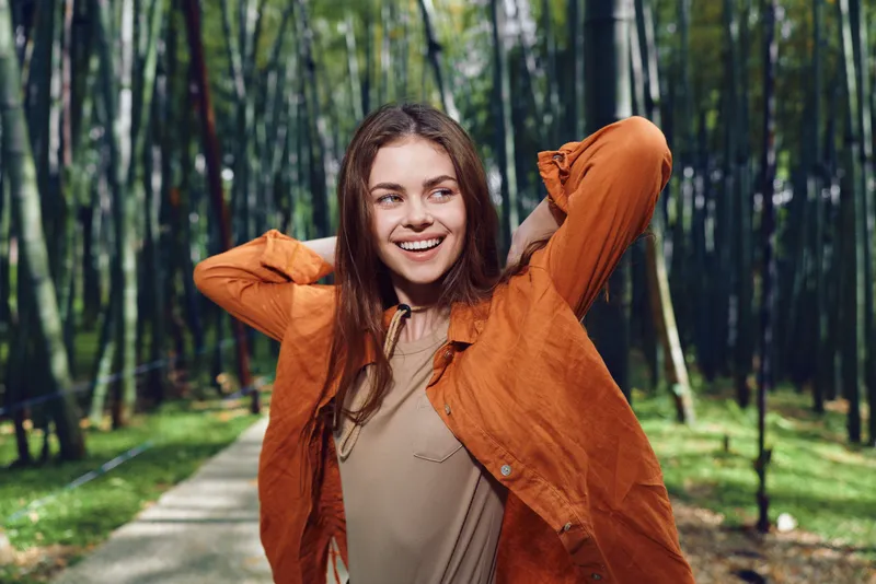Woman smiling in orange jacket on forest path outdoors, portrait of happy young woman stretching with hands behind head, nature walk and casual autumn lifestyle scene.