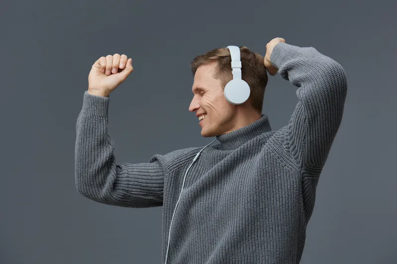 Smiling man wearing headphones enjoying music, raising fist in celebration, casual gray sweater, studio portrait on plain gray background, positive mood concept.