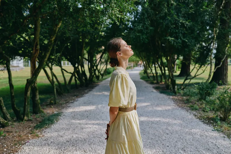 woman dress park path outdoor nature sunlight serene gaze as she stands on a gravel lane in a lush park looking upward, creating a peaceful summer mood with soft light through trees