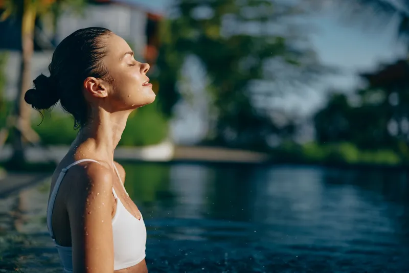 Young woman in white swimsuit relaxing in outdoor pool with eyes closed, enjoying sunlight and fresh air. Concept of wellness, lifestyle, and summer relaxation in natural setting.