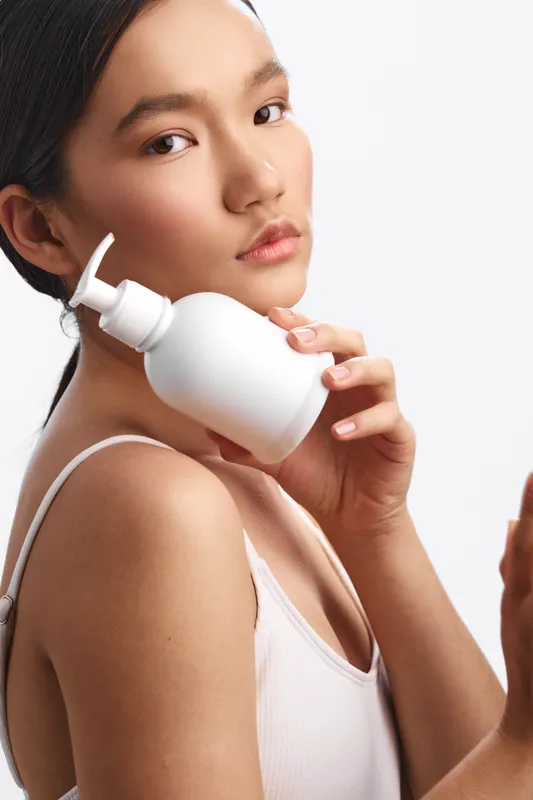 Asian woman with a calm expression applies skincare using a pump bottle in a clean bathroom setting, highlighting a gentle self care routine and healthy skin.