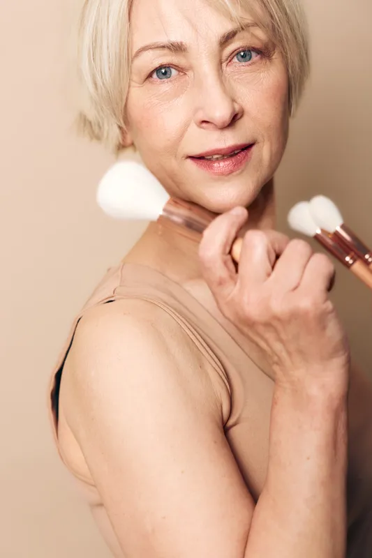 Mature woman with short blonde hair holds makeup brushes close to face, smiling softly against beige background. Concept of beauty, skincare, confidence, and natural aging care.