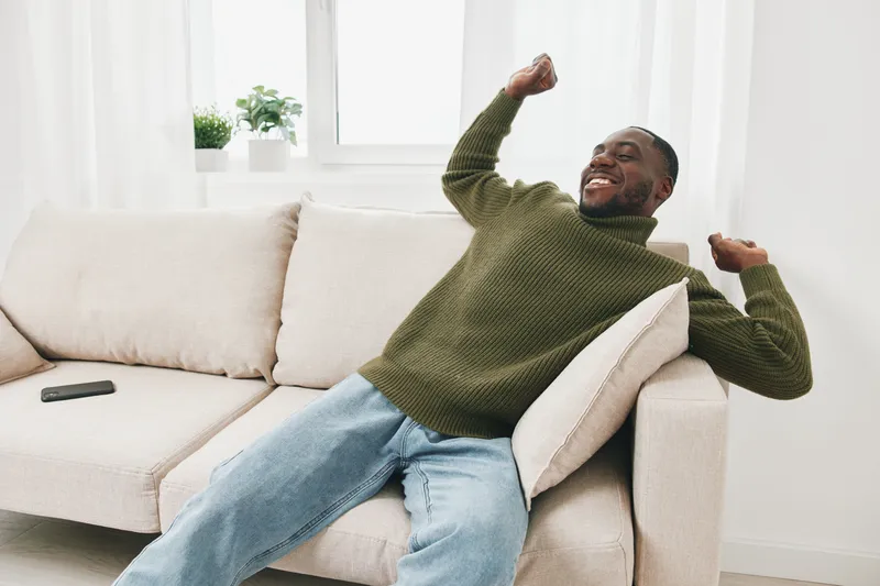 Happy young African American man in cozy sweater expressing joy on sofa at home, symbolizing relaxation and comfort in a bright, modern living room