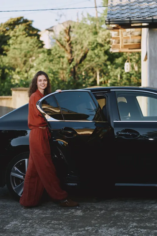Woman lifestyle moment outdoors beside a car, smiling with relaxed stance, sunlit scene, flowing dress, natural colors, lush greenery in the background, and a subtle breeze