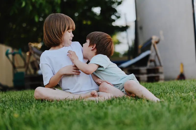 Two children enjoying a playful moment outdoors, sitting on the grass with smiles, conveying happiness and innocence in a bright setting.