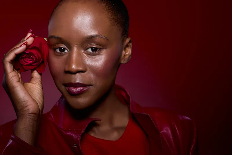 Close-up of confident young black woman with dark skin holding red rose near face against red background. Concept of beauty, elegance, and floral romance in studio portrait.