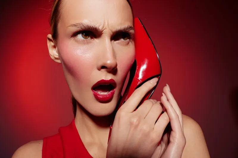 Woman in red dress holds red high heel shoe to her face with surprised expression on red background. Concept of fashion, beauty, and bold style.