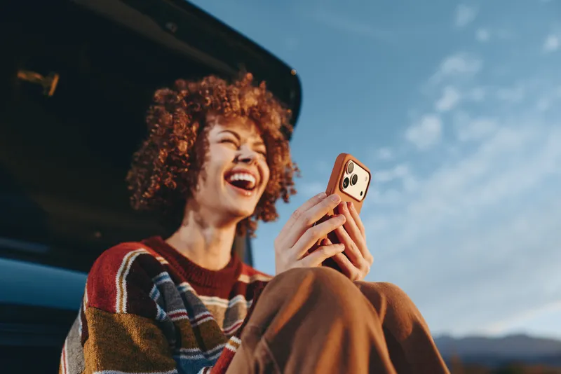 Woman with curly hair smiling while taking a selfie with smartphone near a parked car outdoors, lifestyle moment, soft daylight, blue sky background, joyful travel summer scene