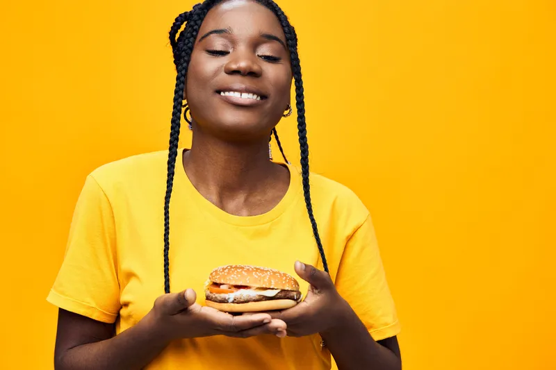 Joyful woman with braids holding a delicious sandwich against a vibrant yellow background, showcasing a healthy eating concept and seasonal freshness