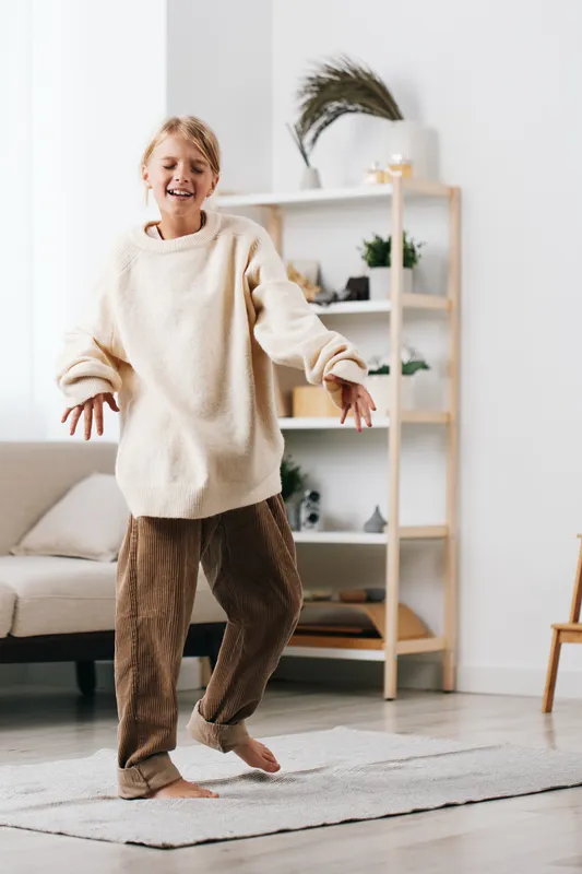 Joyful girl dancing in a cozy living room, wearing a chunky cream sweater and corduroy pants, captured in a bright, warm home setting with soft natural light.