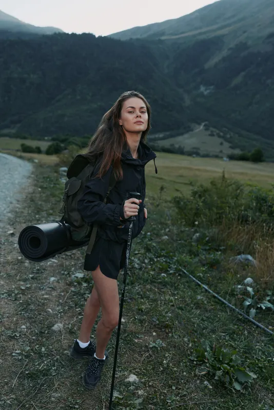 A confident hiker stands on a trail beside a river in a rugged mountain valley. She wears a backpack and carries a rolled mat, exuding independence and outdoor spirit.