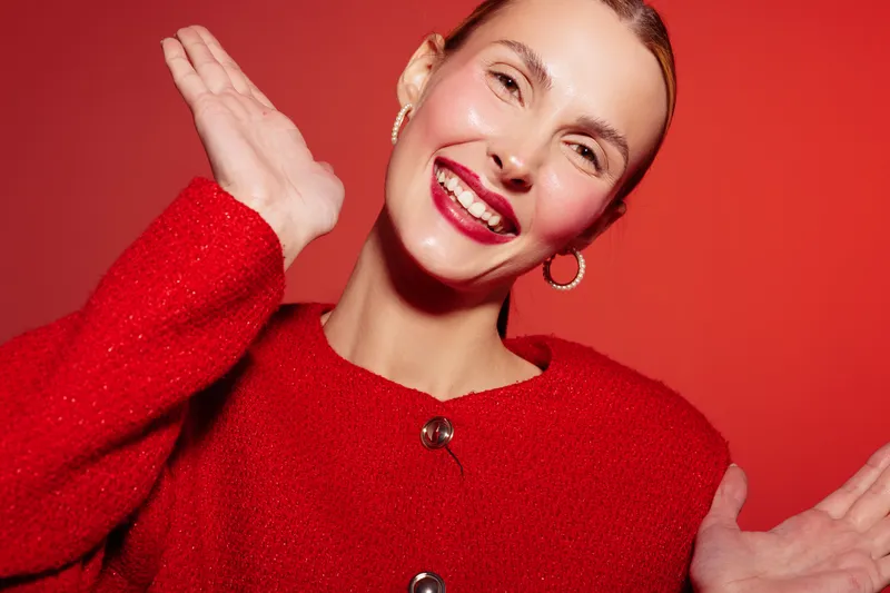 Smiling woman in red sweater poses with hands raised on red background. Bright makeup and hoop earrings create cheerful and confident mood in studio portrait.