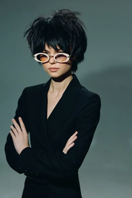 Stylish woman with short black hair in white sunglasses and black blazer posing confidently against gray background