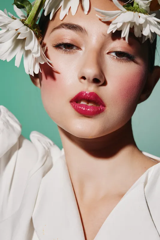 Close-up of a young woman with glossy red lips and dewy skin wearing white daisy petals as an accessory, showcasing beauty creativity and fashion styling on green background.