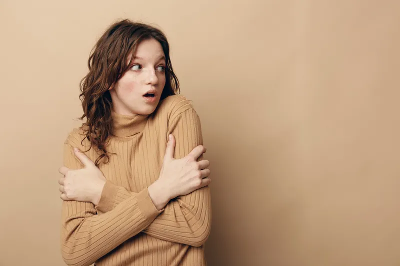 Surprised young woman in brown sweater hugs herself and looks aside with wide eyes on beige background. Concept of shock, fear, cold, and unexpected emotions in casual style.