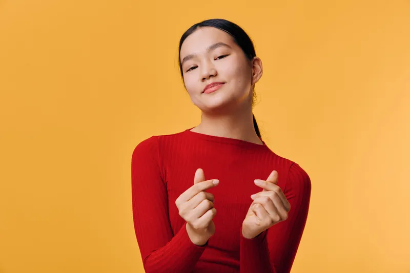 Young woman wearing red sweater, posing with finger heart gesture, smiling confidently on yellow background for positive emotions and casual portrait.