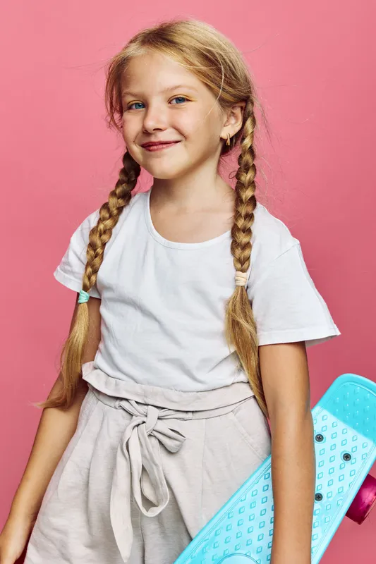 Smiling girl with braided hair holds blue skateboard standing against pink background. Casual outfit with white t-shirt and beige shorts. Youth leisure concept.