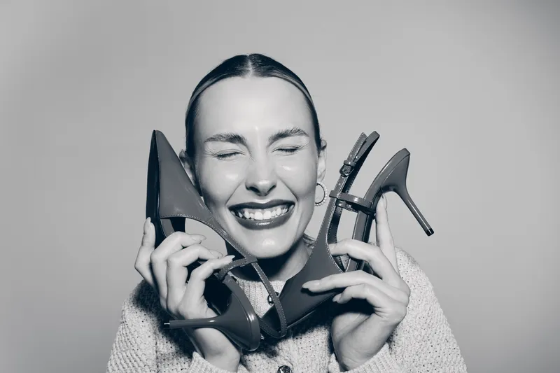 Happy Caucasian woman with fair skin smiling with closed eyes holds two high heel shoes near face. Concept of fashion, joy, and shoe shopping.