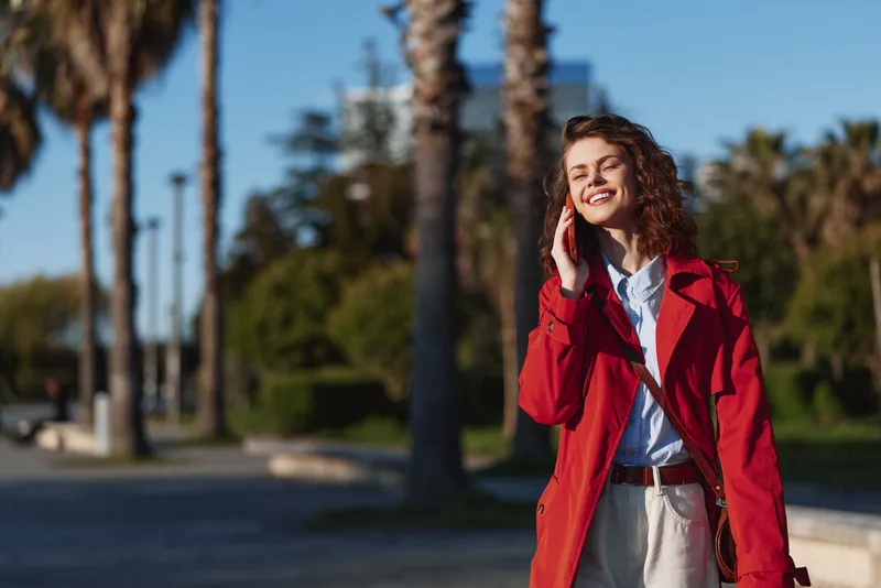 Smiling woman in red coat talking on smartphone outdoors on sunny day with palm trees in the background. Casual style, communication, happiness, urban lifestyle concept.