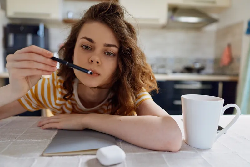 A young woman rests her chin on a notebook while holding a pen in a bright home kitchen, conveying a focused study moment and quiet concentration.
