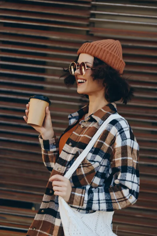 Woman holding coffee cup, smiling in autumn beanie and sunglasses on urban street, candid lifestyle portrait with authenticity, golden hour glow, mindful living and emotional storytelling.