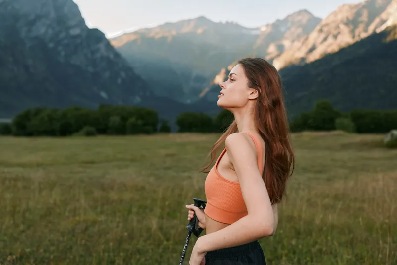 A serene woman stands in a grassy meadow with distant mountains and a clear sky, wearing an orange top. She gazes into the distance, exuding calm, freedom, and a zest for outdoor adventure.