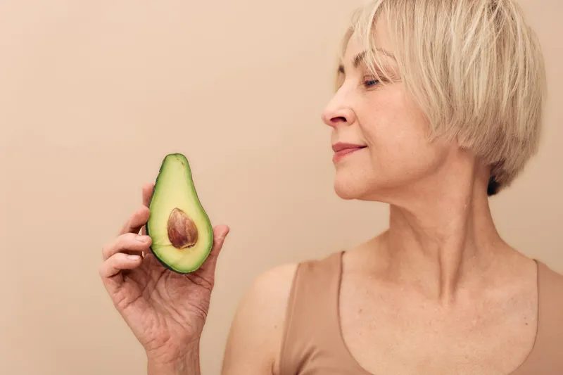 Mature woman with short blonde hair holds half an avocado close to her face on beige background. Concept of healthy eating, natural skincare, nutrition, and wellness for seniors.